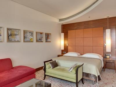 Camera d'hotel moderna con letto matrimoniale, panca, divano rosso e quadri appesi.