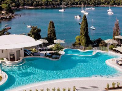 Großer Hotelpool mit Sonnenschirmen, Liegestühlen und Blick auf das Meer mit segelnden Booten.