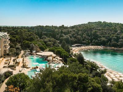 Hotel mit Pool und Sonnenliegen neben einem bewaldeten Strand mit klarem Wasser.