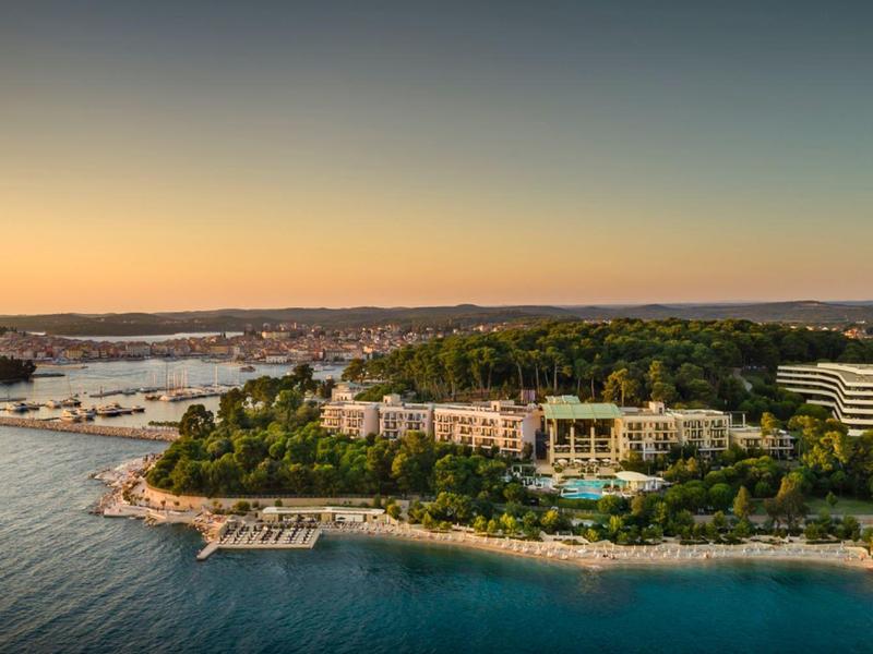 Vista panoramica di un resort sul mare con spiaggia, pineta e porto al tramonto.