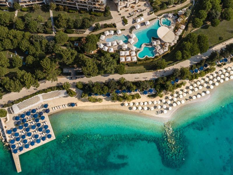 Veduta aerea di una spiaggia con ombrelloni, piscina e mare turchese.