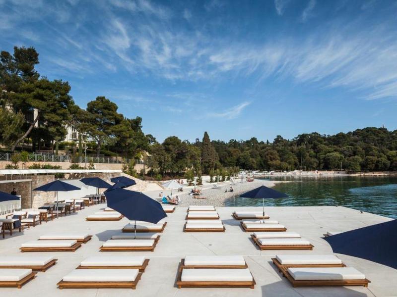 Spiaggia con lettini bianchi e ombrelloni blu davanti a un paesaggio boscoso e cielo limpido.