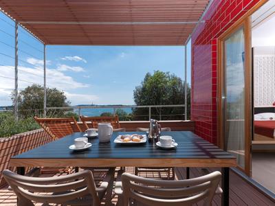 Balkon mit Tisch, Stühlen und Blick auf das Meer, angrenzend an ein Hotelzimmer mit roter Wand.