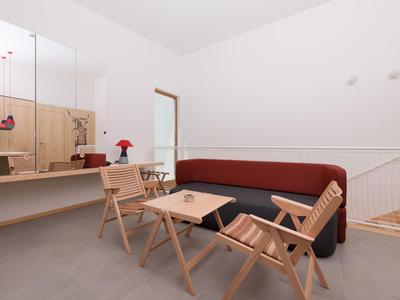 Minimalistisch eingerichteter Hotel-Wartebereich mit Holzstühlen, Tisch und rotem Sofa vor weißer Wand.