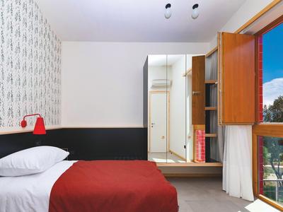 Hell eingerichtetes Hotelzimmer mit Doppelbett, roter Bettdecke, Fenster mit Blick auf Balkon und blauen Himmel.