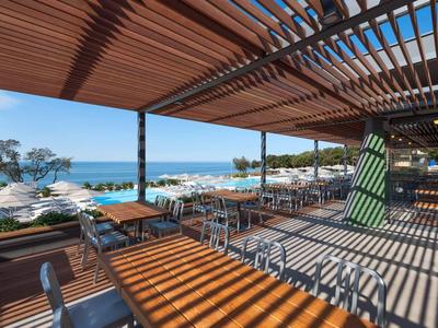 Overdekt terras met tafels en stoelen met uitzicht op zee en zwembad in de zon.