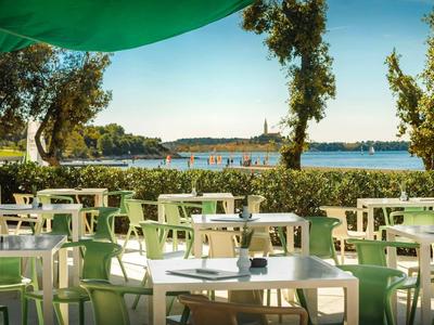 Buitenrestaurant met witte tafels en stoelen, uitzicht op zee en bomen op de achtergrond.