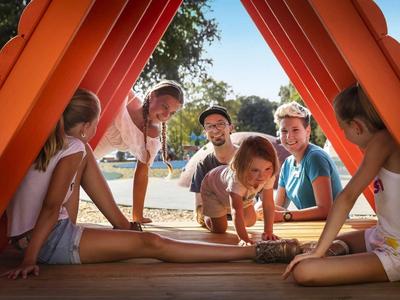 Dzieci bawią się na zewnątrz pod czerwoną drewnianą konstrukcją w słoneczny dzień.