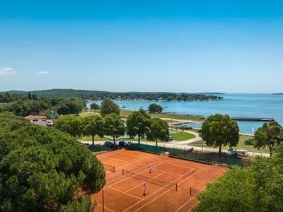 Tennisplätze vor einem blauen See mit Bäumen und leicht bewölktem Himmel.