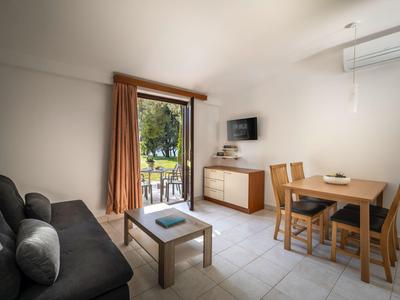Helles Hotelzimmer mit Sofa, Esstisch, TV und Balkon mit Blick auf Bäume.