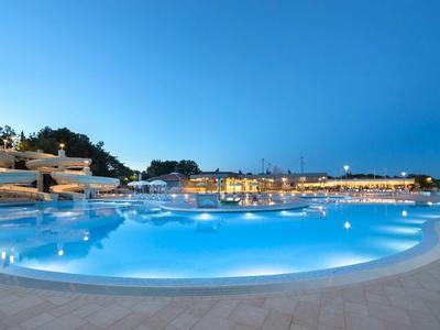 Großer beleuchteter Pool mit klarem Wasser, umgeben von hellen Gebäuden und blauem Nachthimmel.