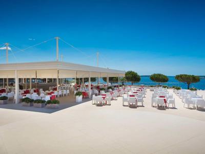 Duży otwarty taras z białymi stolikami i krzesłami przed restauracją z widokiem na niebieskie morze.