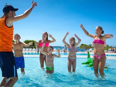 Kinder und Betreuer tanzen im Pool bei sonnigem Himmel in einem Urlaubsresort.