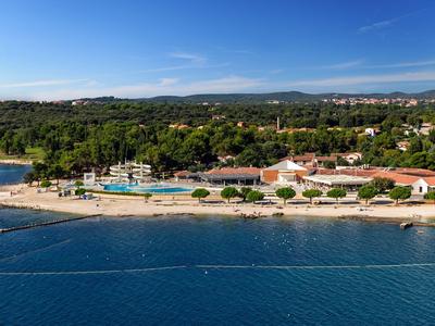 Strandresort mit Pools, Sandstrand und Hotelgebäuden vor bewaldeten Hügeln und blauem Himmel.