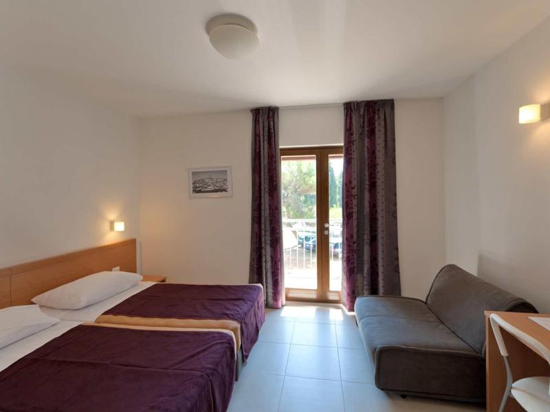 Pokój hotelowy z dwoma pojedynczymi łóżkami, sofą, biurkiem i dostępem do balkonu przez szklane drzwi.