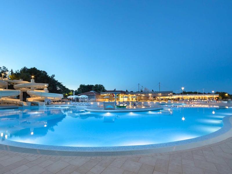 Duży oświetlony basen hotelowy nocą z jasnym niebieskim niebem w tle.