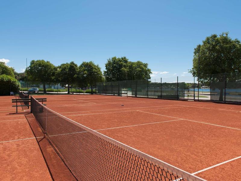 Otwarte, ceglaste boisko do tenisa z czystym niebem i drzewami w tle.