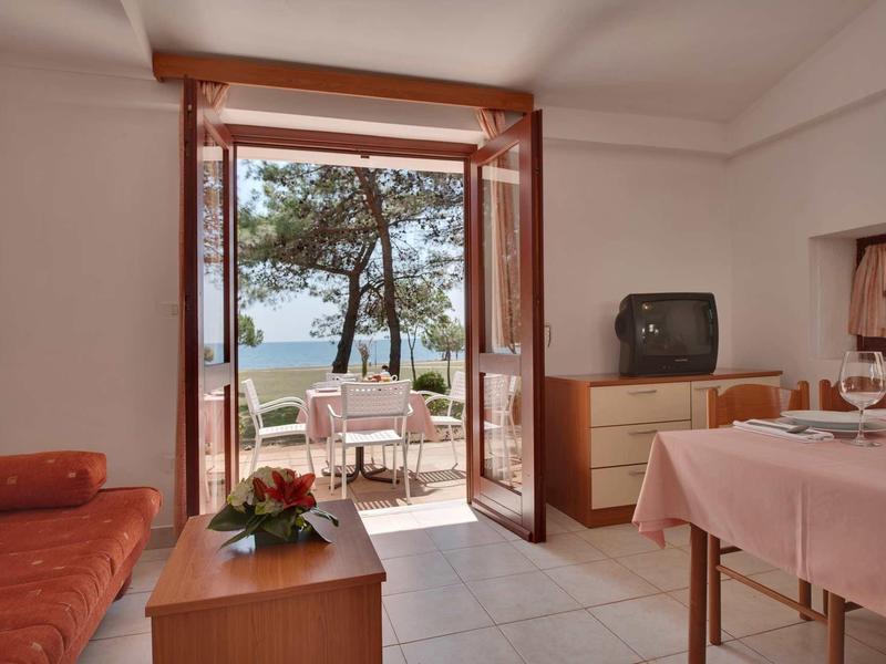 Przytulny pokój hotelowy ze stołem, telewizorem i balkonem z widokiem na morze i drzewa.