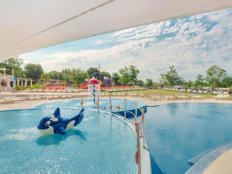 Groot modern buitenzwembad met waterfonteinen en liggedeelte onder blauwe lucht.