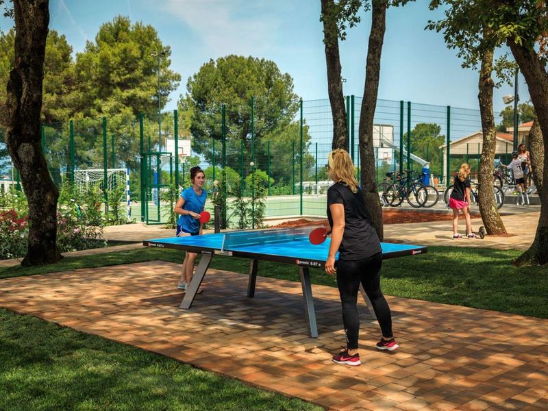 Twee mensen spelen tafeltennis aan een buitentafel naast tennisbanen in een park.