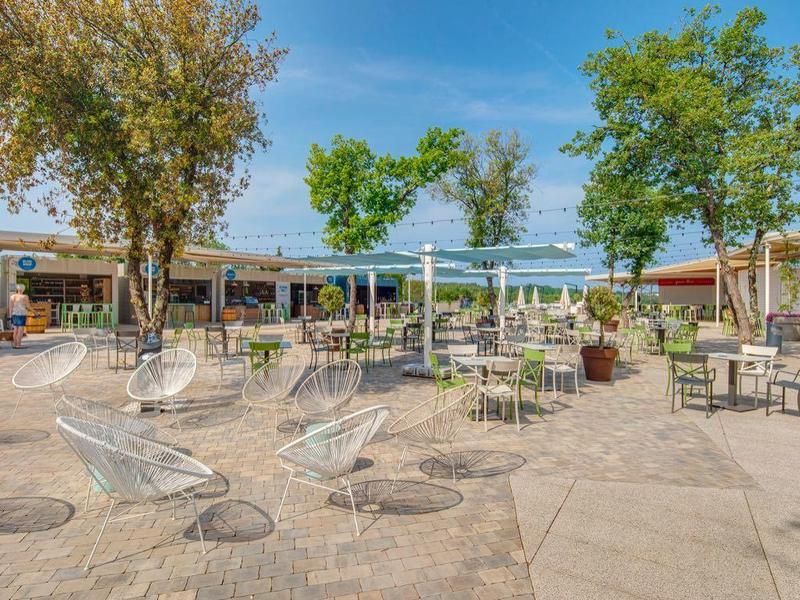 Ruim hotelterras met tafels, stoelen en grote bomen onder een blauwe lucht.