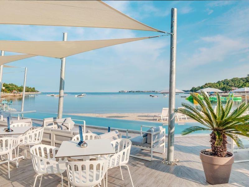 Terras met witte tafels en stoelen aan het water met zonneschermen en planten.