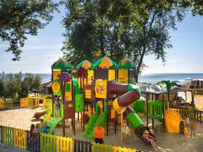 Área de juegos infantil colorida con toboganes y estructuras de escalada en la playa bajo árboles.