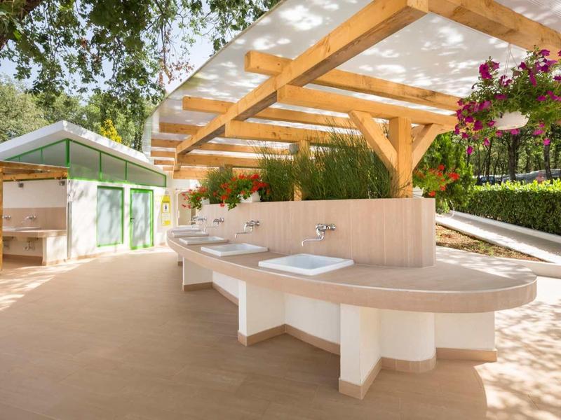 Gran terraza con techo de madera, mesas y bancos, rodeada de árboles y flores.