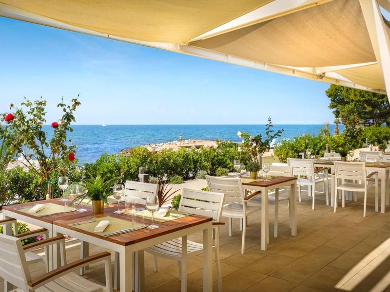 Terraza luminosa con mesas y sillas blancas con vista al mar y plantas florecientes.