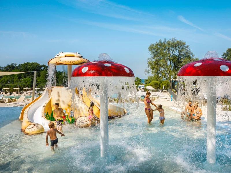 Parque acuático con fuentes en forma de seta y tobogán de agua bajo un clima soleado.