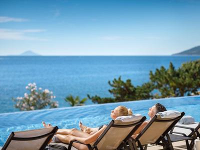 Zwei Personen entspannen auf Liegestühlen am Pool mit Meerblick und blauem Himmel.