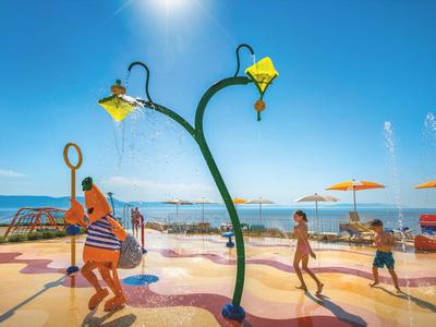 Niños y adultos jugando en una fuente con luz de farolas en un día soleado junto a la playa.