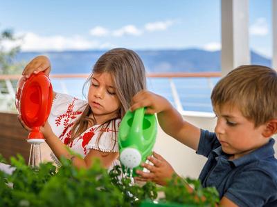 Niños regando plantas verdes en un balcón con vistas al mar y cielo azul despejado.