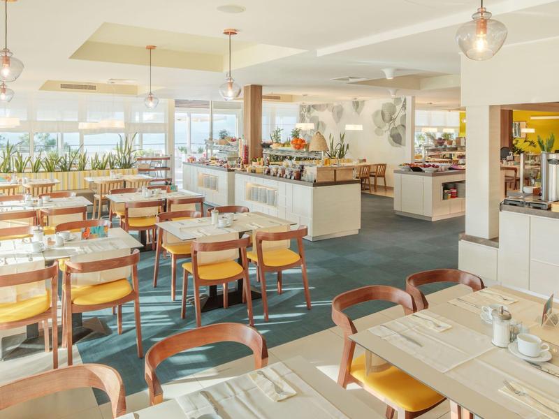 Sala de desayuno luminosa con mesas y sillas de madera, ventanas grandes y área de buffet.