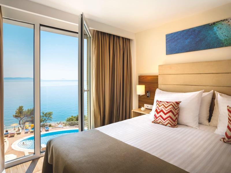 Habitación de hotel luminosa con vista al mar, ventana abierta, cama con cojines decorativos y piscina afuera.