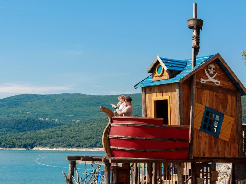 Niño juega en una casa de juegos con forma de barco pirata con vista a montañas y mar.