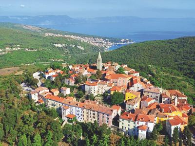 Vista aérea de un pintoresco pueblo costero con techos rojos, rodeado de colinas verdes y vista al mar.