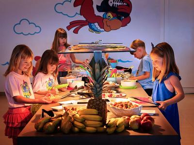 Niños comiendo y sirviéndose en una mesa larga con plátanos y piña en una sala colorida.