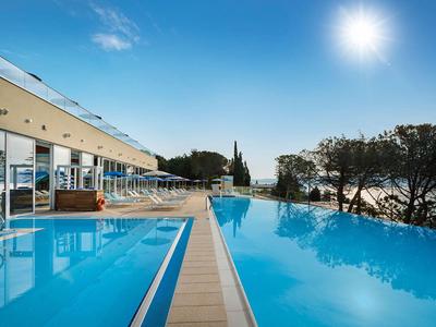 Basen hotelowy z tarasem i widokiem na morze pod jasnym niebieskim niebem i słońcem.