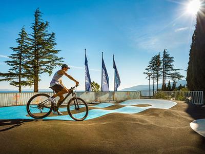 Osoba jedzie na rowerze ścieżką obok basenu z widokiem na morze i flagi w tle.