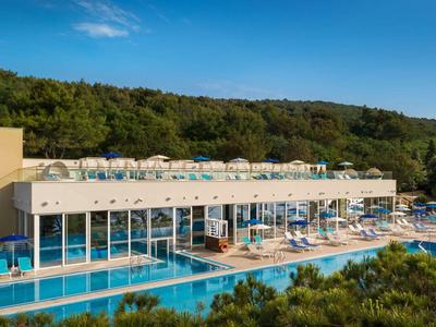 Modernes Gebäude mit großen Glasfenstern, integriertem Pool und vielen Sonnenschirmen, umgeben von Wald.