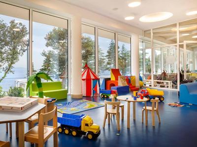 Sala de juegos infantil luminosa con juguetes, mesas y vistas al exterior con árboles y mar.