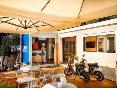 Przytulny taras na zewnątrz z białymi krzesłami, skuterami elektrycznymi i wejściem do sklepu pod dużymi parasolami.