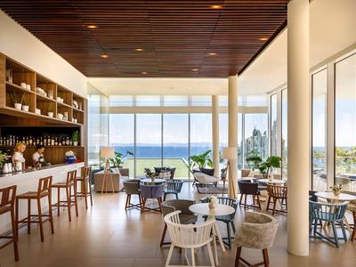 Interior moderno de un bar con sillas y mesas frente a grandes ventanales que ofrecen vista al mar.