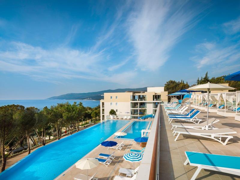 Piscina moderna de hotel con tumbonas azules y vista al mar bajo cielo despejado.