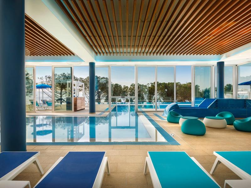 Área moderna de piscina interior con tumbonas azules, pufs y grandes ventanas con vista al exterior.