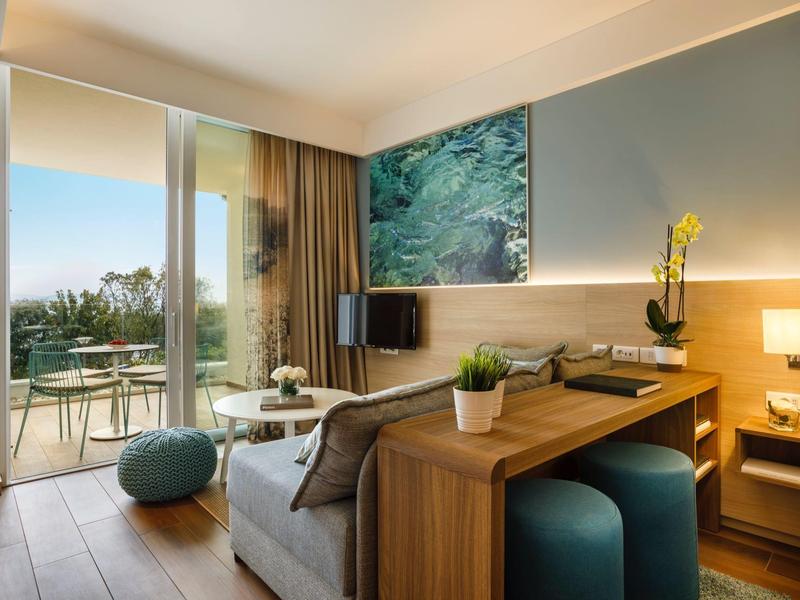 Habitación de hotel moderna con balcón, sofá, escritorio de madera y vista al verde.