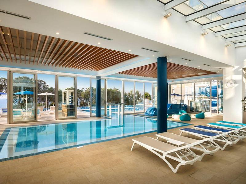 Piscina interior con tumbonas blancas y grandes ventanales que muestran un paisaje exterior soleado.