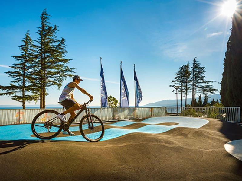 Mujer monta bicicleta en una pista con banderas y árboles bajo el sol junto al mar.
