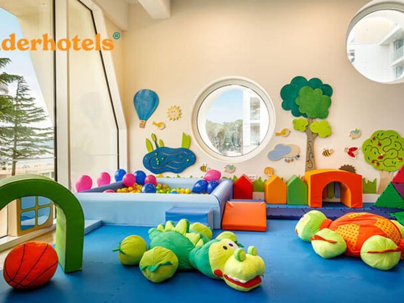 Área de juegos colorida con juguetes de felpa, almohadones y ventanas redondas en hotel familiar.